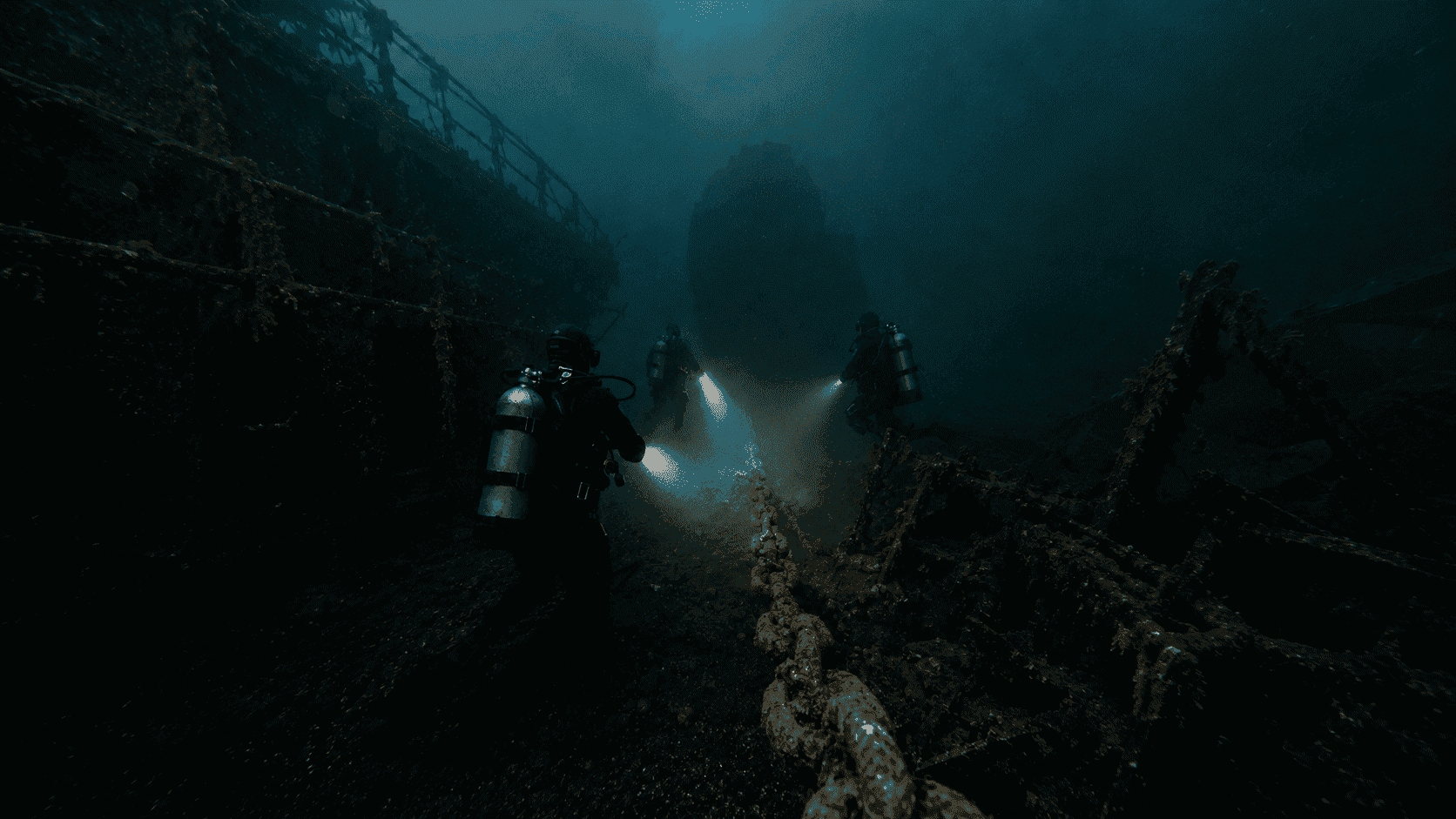 Scuba divers following the rusted chain underwater toward a sunken vessel