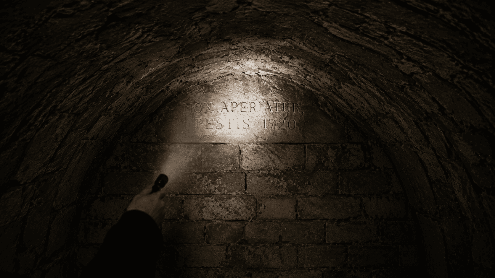A flashlight illuminating a carved Latin inscription reading NON APERIATUR PESTIS 1720 above a sealed brick wall