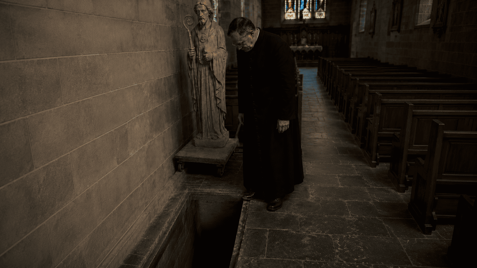 Father William standing over a dark opening in the church floor beneath a St. Benedict statue