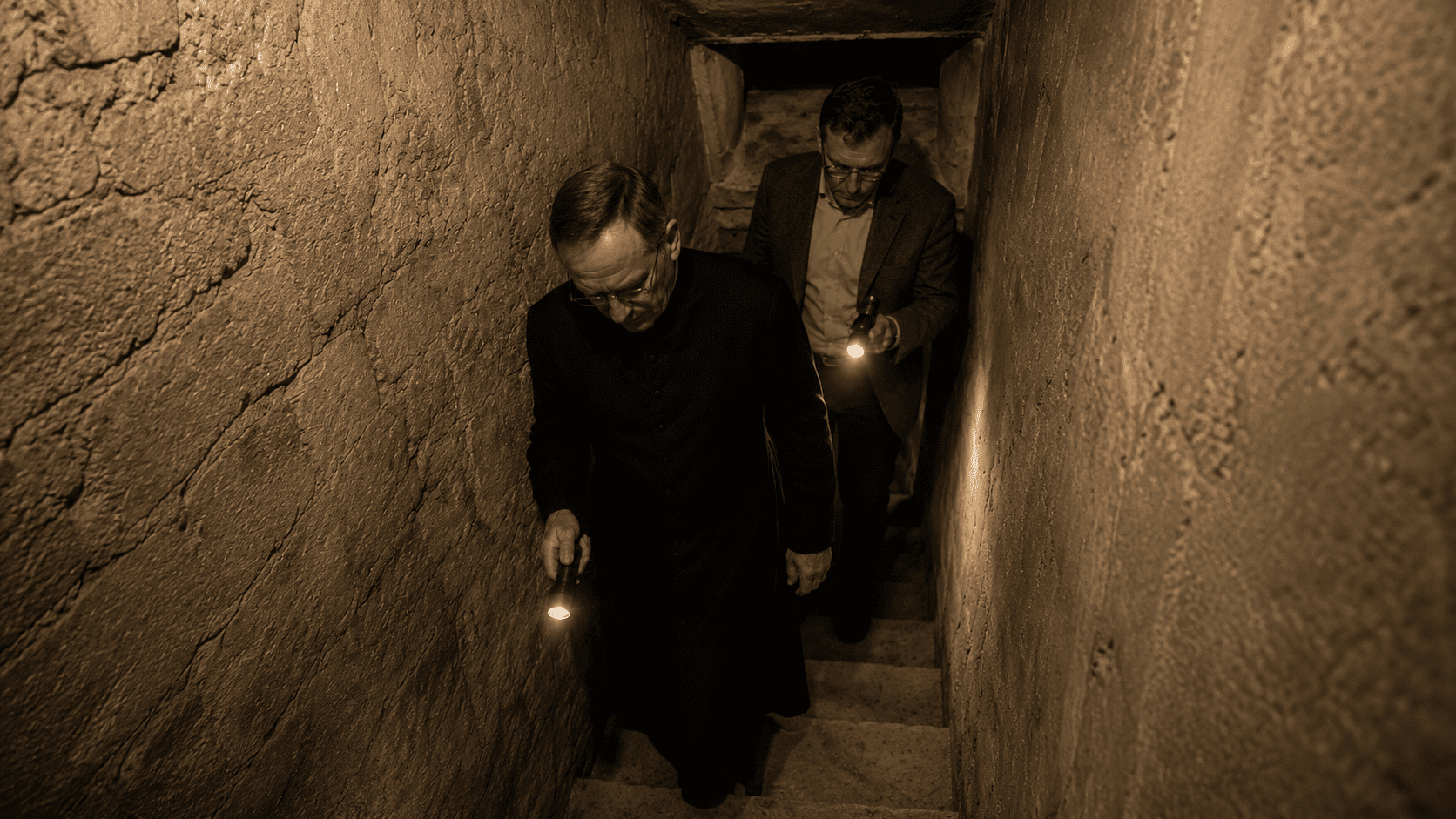 Father William and Dr. Arthur Vance descending a narrow stone staircase underground with flashlights